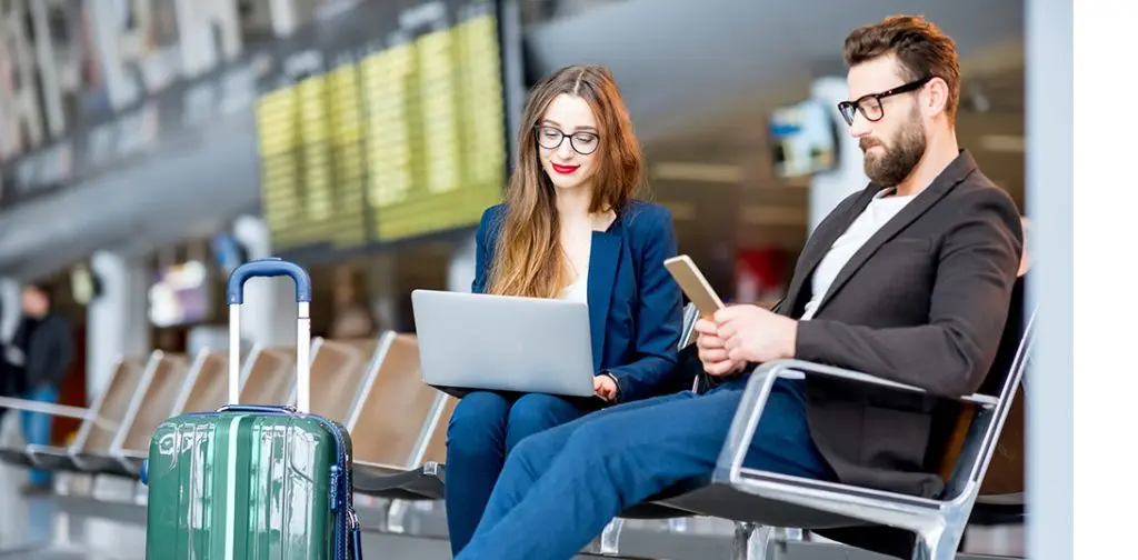 دو مسافر در فرودگاه که یکی در حال استفاده از لپ‌تاپ و دیگری با موبایل، نشان‌دهنده‌ی دسترسی به اینترنت بانک از خارج کشور در حین سفر.